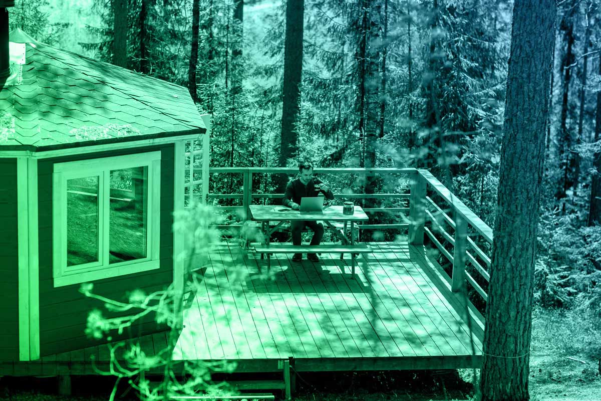 middle-aged man with laptop sitting down at a wooden table on country house terrace located among trees in a woodland